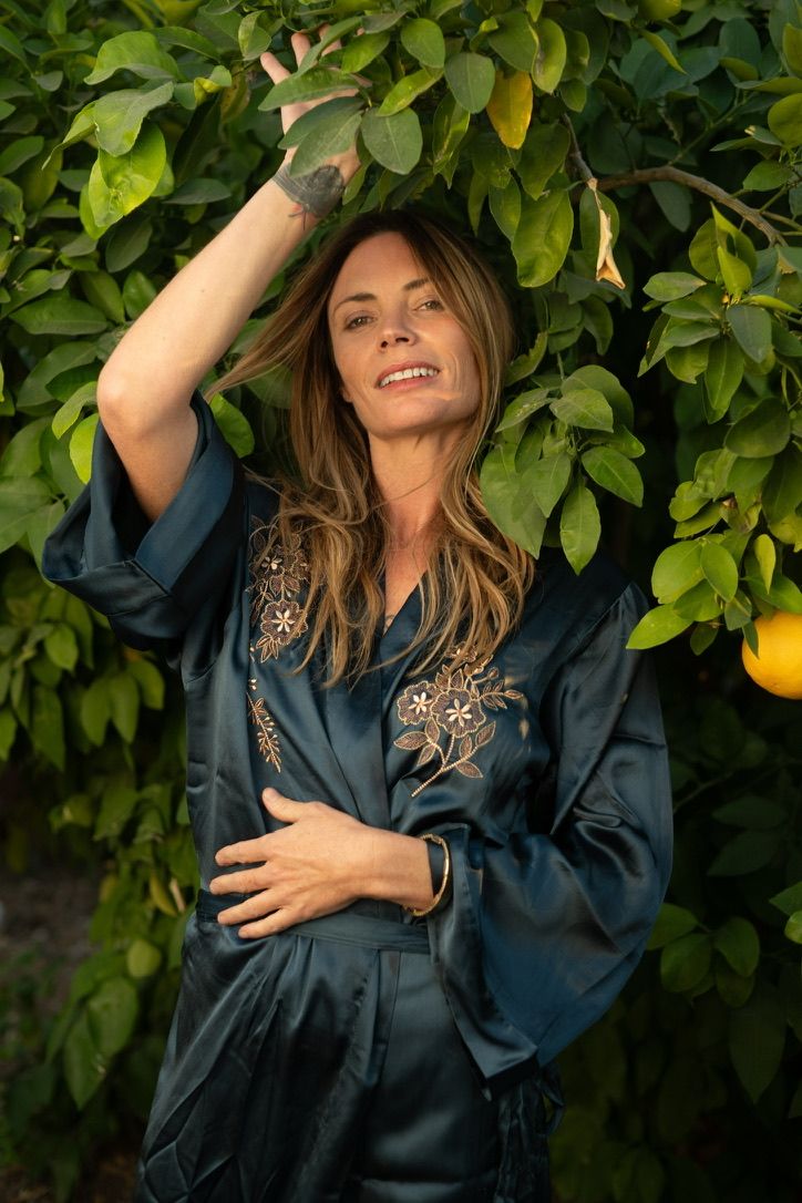 Rachel smiling in a dark satin robe beneath a citrus tree